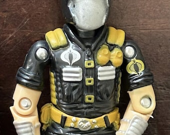 May include: A plastic action figure wearing a black helmet with silver goggles and a face mask. The figure has a black vest with yellow accents, silver details, and a Cobra logo on the chest and left sleeve. It features light-colored arms with silver wrist guards.
