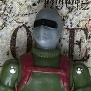 May include: A green and red action figure with a gray face mask and black visor. The figure is wearing a green jumpsuit with a red harness.