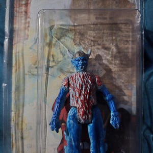 May include: A blue action figure with red and white accents, packaged in a clear plastic case. The figure has a horned helmet and a detailed torso. The packaging features a Coca-Cola logo and vintage-style graphics.