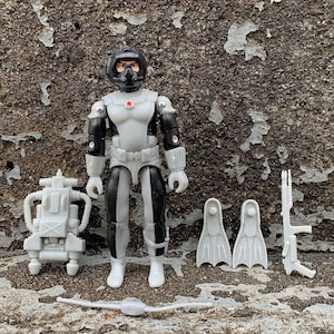 Puede incluir: Una figura de acción en blanco y negro con un casco de buceo y una luz roja en el pecho. La figura sostiene un arma de fuego en blanco y negro y lleva una mochila en blanco y negro. La figura también lleva aletas en blanco y negro y un aparato respiratorio en blanco y negro.