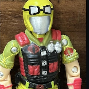 May include: A vintage action figure of a character in a yellow helmet with silver goggles and a silver face mask. The figure wears a green and black uniform with red accents and a Cobra logo on the chest.