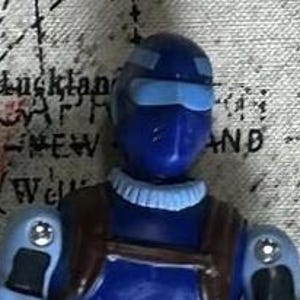 May include: A blue action figure with a light blue helmet and a brown backpack. The figure has a light blue collar and light blue accents on the arms and legs.