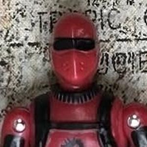 May include: A red action figure with a black helmet and black accents. The figure is wearing a red suit with black details.