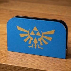 May include: A blue case with a gold Triforce emblem, a symbol from the Legend of Zelda games. The case has a rectangular shape with curved edges. It is placed on a wooden surface.