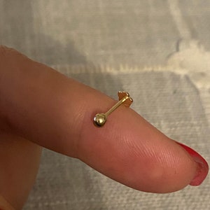 Pode incluir: Um pequeno brinco dourado com uma bola numa extremidade e um design em forma de flor na outra. O brinco é mostrado num dedo, destacando o seu tamanho e design delicados. O brinco é provavelmente para um piercing na cartilagem.