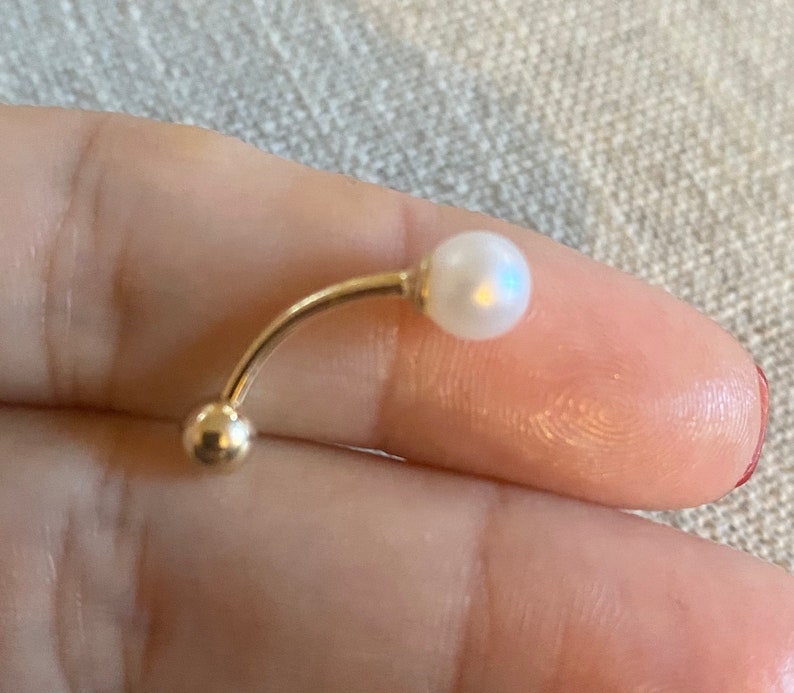Op de afbeelding: Een gebogen, goudkleurige navelpiercing met een kleine, witte parel aan &eacute;&eacute;n uiteinde en een gouden bal aan het andere. Het sieraad wordt in een hand gehouden, wat het delicate ontwerp benadrukt.