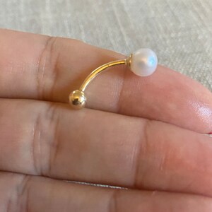 Op de afbeelding: Een gebogen, goudkleurige navelpiercing met een kleine, ronde gouden bal aan &eacute;&eacute;n uiteinde en een witte parel aan de andere kant. De sieraden worden getoond tegen een neutrale achtergrond.