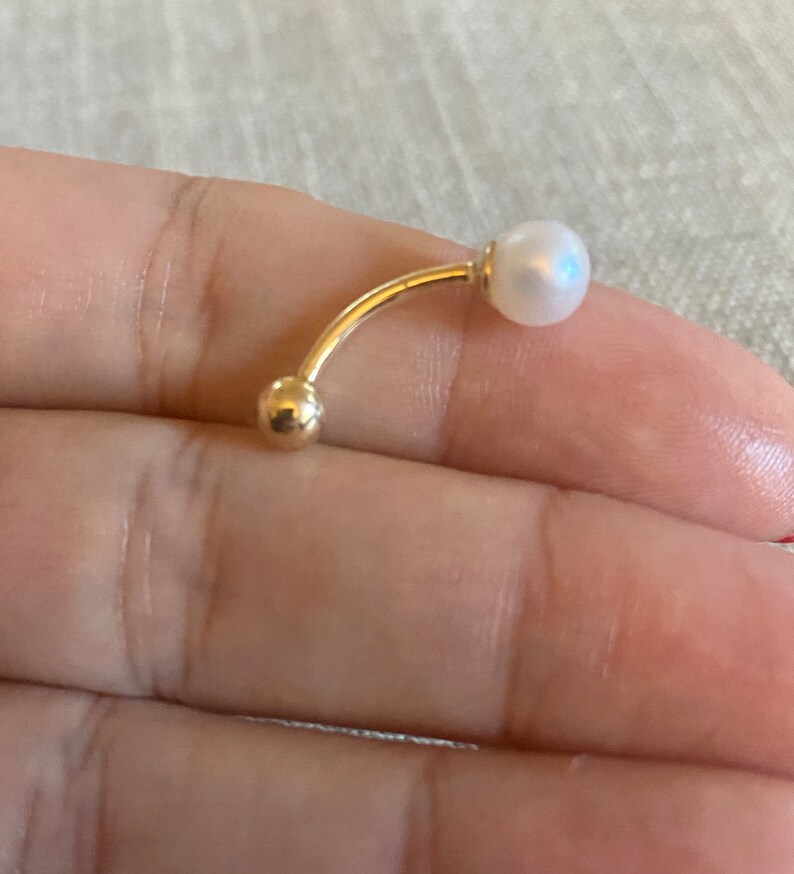 Op de afbeelding: Een gebogen, goudkleurige navelpiercing met een kleine, ronde gouden bal aan &eacute;&eacute;n uiteinde en een witte parel aan de andere kant. De sieraden worden getoond tegen een neutrale achtergrond.