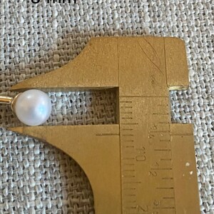 Op de afbeelding: Een witte parel op een gouden oorbel, gemeten met een messing schuifmaat. De parel heeft een diameter van ongeveer 8 mm. De oorbel staat tegen een getextureerde, lichtgrijze achtergrond.