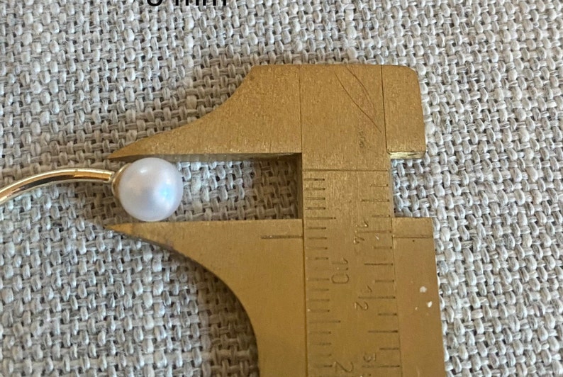 Op de afbeelding: Een witte parel op een gouden oorbel, gemeten met een messing schuifmaat. De parel heeft een diameter van ongeveer 8 mm. De oorbel staat tegen een getextureerde, lichtgrijze achtergrond.