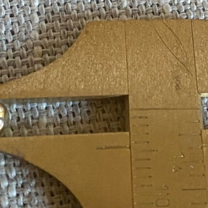 Op de afbeelding: Een goudkleurige metalen schuifmaat meet een klein, glanzend, bolvormig object. Het object lijkt een kraal of een kleine bal te zijn. De schuifmaat is gemarkeerd met metingen in centimeters.