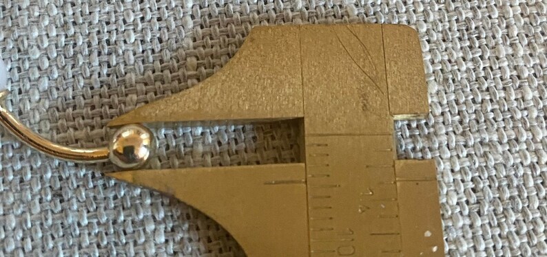 Op de afbeelding: Een goudkleurige metalen schuifmaat meet een klein, glanzend, bolvormig object. Het object lijkt een kraal of een kleine bal te zijn. De schuifmaat is gemarkeerd met metingen in centimeters.
