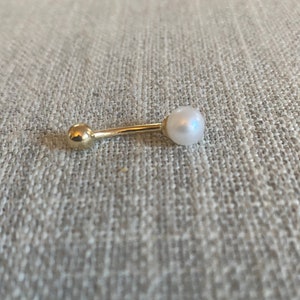 Op de afbeelding: Een goudkleurige, rechte oorbel in halterstijl met een witte parel aan &eacute;&eacute;n uiteinde en een kleine gouden bal aan de andere kant. De oorbel ligt op een neutraal gekleurd, gestructureerd oppervlak.