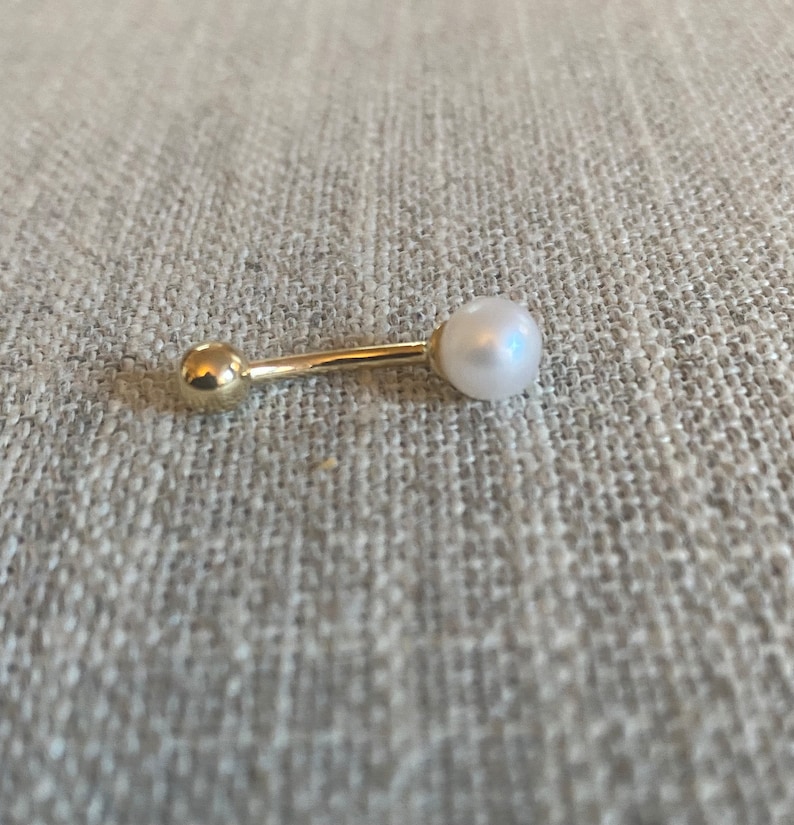 Op de afbeelding: Een goudkleurige, rechte oorbel in halterstijl met een witte parel aan &eacute;&eacute;n uiteinde en een kleine gouden bal aan de andere kant. De oorbel ligt op een neutraal gekleurd, gestructureerd oppervlak.