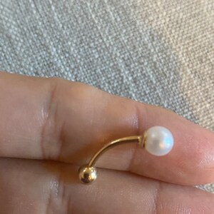 Op de afbeelding: Een gebogen navelpiercing met een goudkleurige metalen staaf en een kleine, ronde gouden bal aan &eacute;&eacute;n uiteinde. Het andere uiteinde heeft een witte, ronde parel. De sieraden worden getoond tegen een neutrale achtergrond.