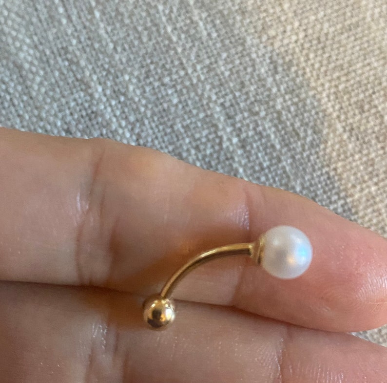 Op de afbeelding: Een gebogen navelpiercing met een goudkleurige metalen staaf en een kleine, ronde gouden bal aan &eacute;&eacute;n uiteinde. Het andere uiteinde heeft een witte, ronde parel. De sieraden worden getoond tegen een neutrale achtergrond.