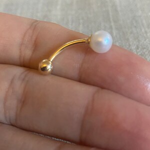 Op de afbeelding: Een gebogen, goudkleurige navelpiercing met een kleine, glanzende witte parel aan &eacute;&eacute;n uiteinde en een gouden bal aan het andere. Het sieraad wordt in de hand van een persoon gehouden en toont het elegante ontwerp en de delicate details.