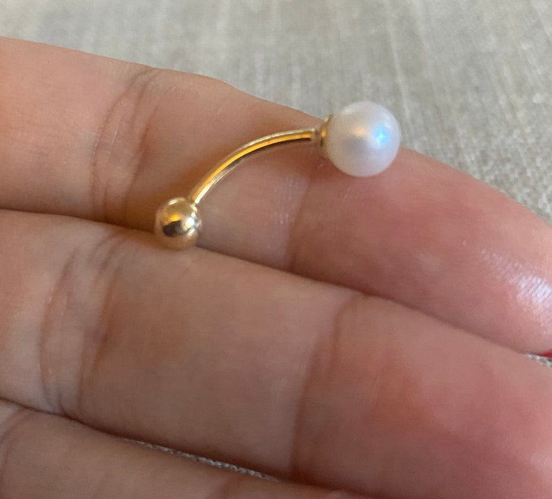 Op de afbeelding: Een gebogen, goudkleurige navelpiercing met een kleine, glanzende witte parel aan &eacute;&eacute;n uiteinde en een gouden bal aan het andere. Het sieraad wordt in de hand van een persoon gehouden en toont het elegante ontwerp en de delicate details.