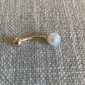 Op de afbeelding: Een goudkleurige navelpiercing met een gebogen staaf. E&eacute;n uiteinde heeft een kleine, ronde gouden bal, de andere een witte, ronde parel. De sieraden zijn geplaatst tegen een neutrale, getextureerde achtergrond.