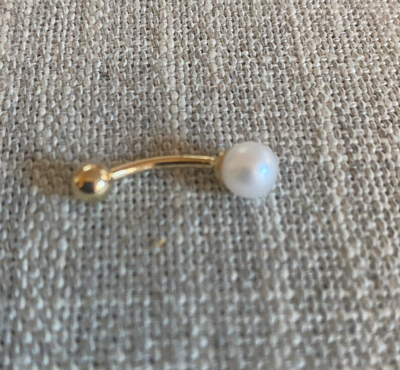Op de afbeelding: Een goudkleurige navelpiercing met een gebogen staaf. E&eacute;n uiteinde heeft een kleine, ronde gouden bal, de andere een witte, ronde parel. De sieraden zijn geplaatst tegen een neutrale, getextureerde achtergrond.