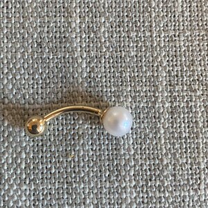 Op de afbeelding: Een goudkleurige, gebogen barbell-piercingsieraden met een kleine gouden bal aan &eacute;&eacute;n uiteinde en een witte parel aan de andere kant. De sieraden zijn geplaatst tegen een getextureerde, neutraal gekleurde achtergrond.