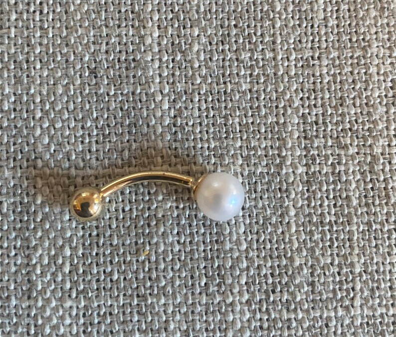 Op de afbeelding: Een goudkleurige, gebogen barbell-piercingsieraden met een kleine gouden bal aan &eacute;&eacute;n uiteinde en een witte parel aan de andere kant. De sieraden zijn geplaatst tegen een getextureerde, neutraal gekleurde achtergrond.