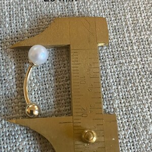 Op de afbeelding: Een goudkleurige gebogen barbell-oorbel met een witte parel aan de bovenkant en een kleine gouden bal aan de onderkant. De oorbel wordt gemeten met een schuifmaat, met een lengte van ongeveer 2,5 cm.