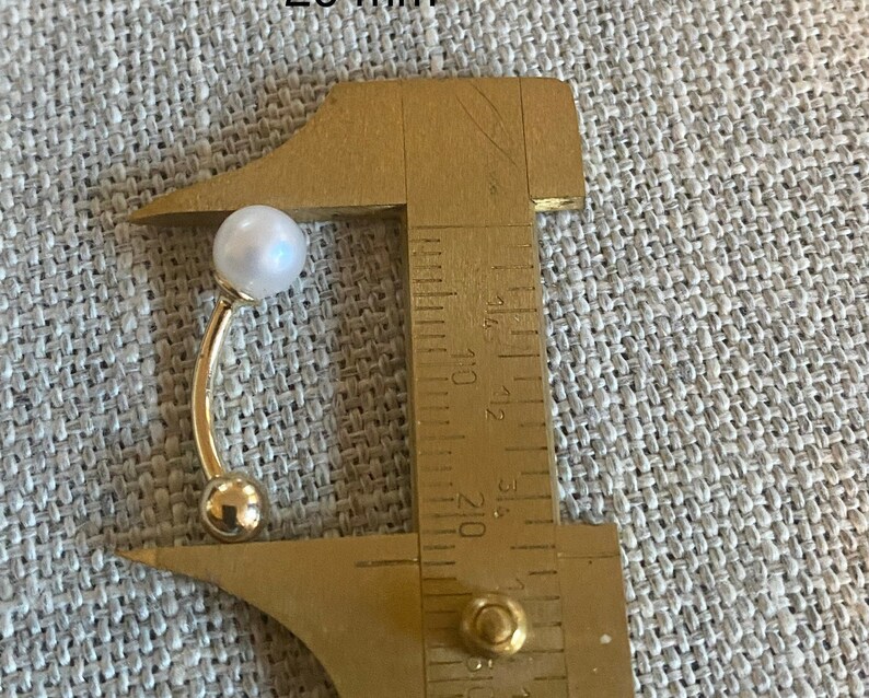 Op de afbeelding: Een goudkleurige gebogen barbell-oorbel met een witte parel aan de bovenkant en een kleine gouden bal aan de onderkant. De oorbel wordt gemeten met een schuifmaat, met een lengte van ongeveer 2,5 cm.