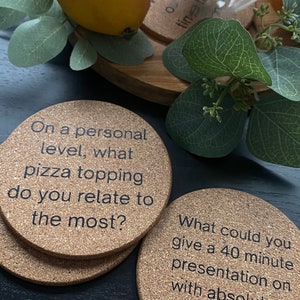 May include: Two round cork coasters with conversation starter questions printed on them. The first coaster says "On a personal level, what pizza topping do you relate to the most?" The second coaster says "What could you give a 40 minute presentation on with absolutely zero preparation?"