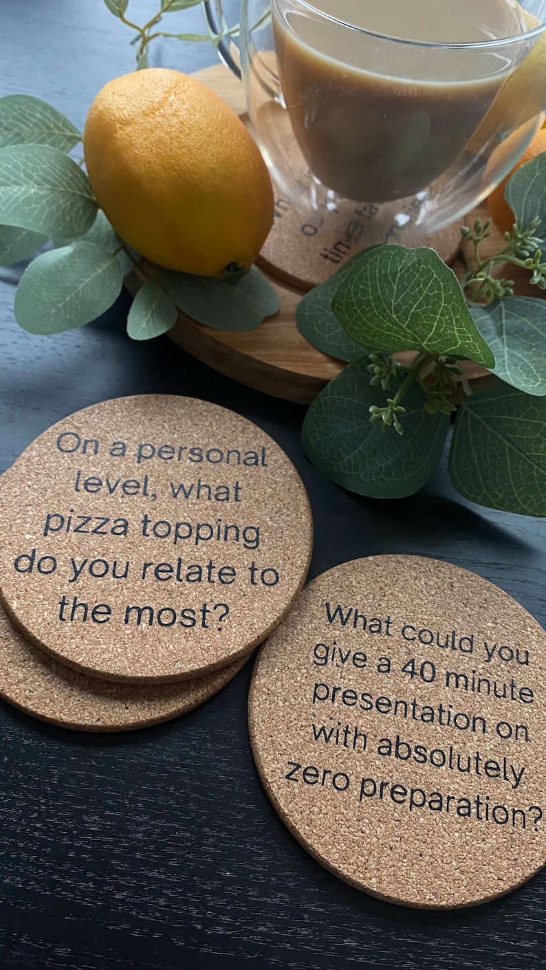 May include: Two round cork coasters with conversation starter questions printed on them. The first coaster says "On a personal level, what pizza topping do you relate to the most?" The second coaster says "What could you give a 40 minute presentation on with absolutely zero preparation?"