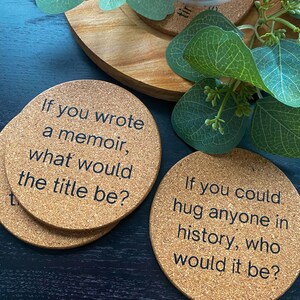 May include: Four cork coasters with black text. The coasters feature conversation starter questions. The first coaster says "If you wrote a memoir, what would the title be?" The second coaster says "If you could hug anyone in history, who would it be?"