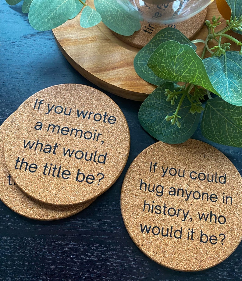 May include: Four cork coasters with black text. The coasters feature conversation starter questions. The first coaster says "If you wrote a memoir, what would the title be?" The second coaster says "If you could hug anyone in history, who would it be?"