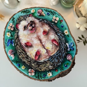 May include: A circular wood slice featuring a hand-painted illustration of a hedgehog lying on its back. The hedgehog is surrounded by a teal background decorated with small white flowers, red and white mushrooms, and blue butterflies.