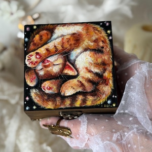 May include: A square wooden box featuring a hand-painted illustration of a ginger tabby cat curled into a ball on a black background. The cat has orange and white fur with dark stripes, and the scene is accented with small, colourful painted stars.