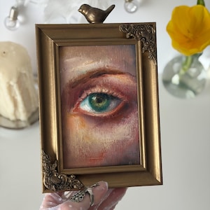 Pode incluir: Uma moldura retangular em tom dourado com um pássaro de metal ornamentado pousado no topo central e detalhes em filigrana nos cantos. A moldura contém uma pintura a óleo de um olho humano com tons de íris verdes e castanhos.
