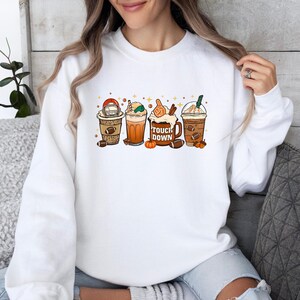 May include: White sweatshirt featuring a graphic of four fall-themed coffee drinks, each with football-related decorations. The phrase "Touch Down" is printed on one of the drinks. The sweatshirt is a crew neck style.