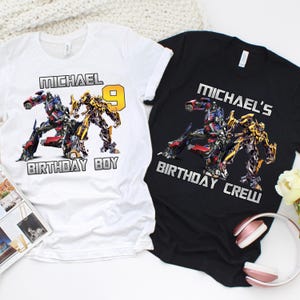 May include: Two t-shirts, one white and one black, featuring a colorful graphic of Transformers characters. The white shirt says "MICHAEL BIRTHDAY BOY" with the number 9. The black shirt says "MICHAEL'S BIRTHDAY CREW".