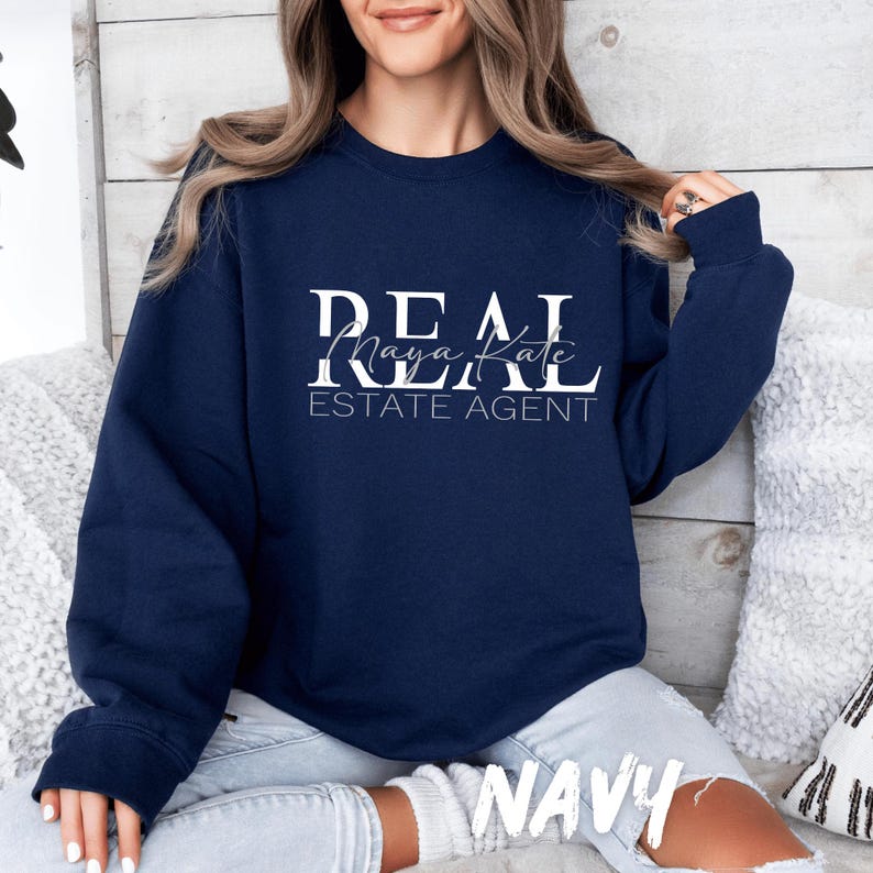 May include: A navy blue crewneck sweatshirt featuring the white text REAL, with the name Maya Kate written in a cursive script font underneath, followed by the text ESTATE AGENT in a clean, sans-serif font centered on the chest.