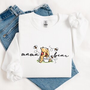 Peut inclure: Sweat-shirt blanc &agrave; manches longues avec un dessin de Winnie l'Ourson tenant un pot de miel, entour&eacute; d'abeilles. Les mots "mama bear" sont &eacute;crits en script. Le sweat-shirt est pos&eacute; sur un jean bleu.