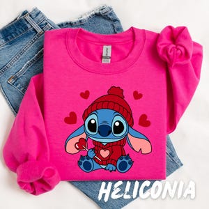 Peut inclure: Un sweat-shirt rose avec un personnage bleu Stitch portant un chapeau et un pull rouge, tenant une sucette rouge. Le personnage est entour&eacute; de c&oelig;urs rouges. Le texte "HELICONIA" est imprim&eacute; en bas du sweat-shirt.
