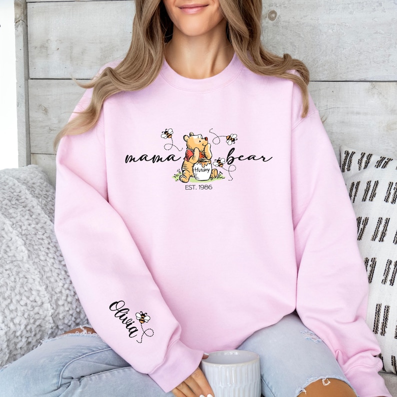 Peut inclure: Un sweat-shirt rose clair avec l'inscription "mama bear" en &eacute;criture cursive, et un dessin de Winnie l'Ourson tenant un pot de miel. Le texte "EST. 1986" est en dessous. Le nom "Olivia" est sur la manche.