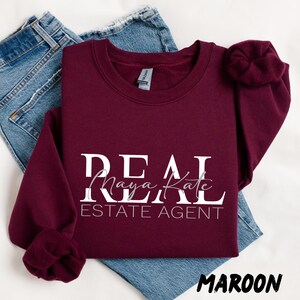 May include: A maroon long-sleeve crewneck sweatshirt featuring the white text REAL ESTATE AGENT with the name Maya Kate in a cursive script font layered over the word REAL. The sweatshirt is displayed on top of a pair of blue denim jeans.