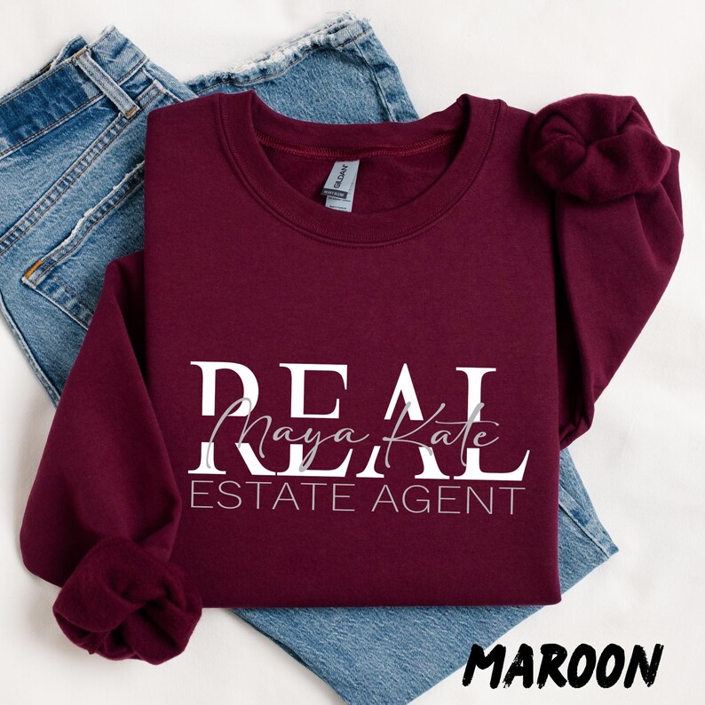 May include: A maroon long-sleeve crewneck sweatshirt featuring the white text REAL ESTATE AGENT with the name Maya Kate in a cursive script font layered over the word REAL. The sweatshirt is displayed on top of a pair of blue denim jeans.