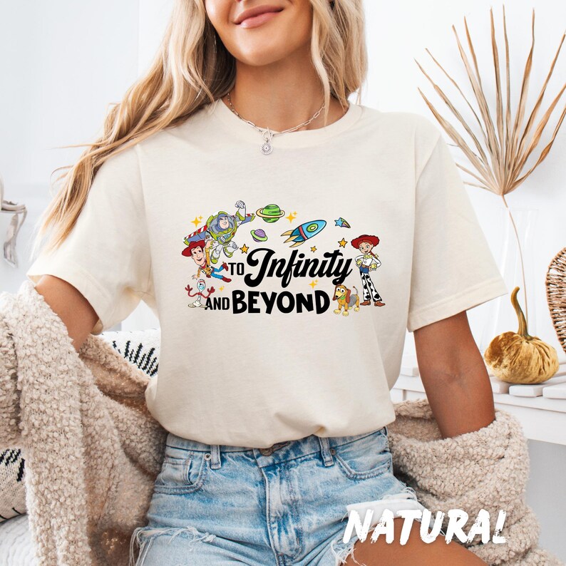 Puede incluir: Una camiseta blanca con un gr&aacute;fico de personajes de la franquicia de pel&iacute;culas Toy Story. El gr&aacute;fico incluye a Woody, Buzz Lightyear y otros personajes. El texto "To Infinity and Beyond" est&aacute; impreso en la camiseta.