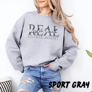 May include: A light gray crewneck sweatshirt featuring the dark gray text REAL ESTATE AGENT in a bold serif font, with the name Leslie Smith printed in a black cursive script across the center of the word REAL.