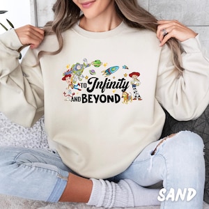 Puede incluir: Una sudadera de color crema con un gr&aacute;fico de personajes de la franquicia de pel&iacute;culas Toy Story. El gr&aacute;fico incluye a Buzz Lightyear, Woody y otros personajes. El texto "To Infinity and Beyond" est&aacute; impreso en la sudadera.