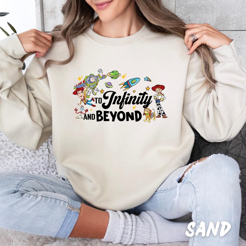 Puede incluir: Una sudadera de color crema con un gr&aacute;fico de personajes de la franquicia de pel&iacute;culas Toy Story. El gr&aacute;fico incluye a Buzz Lightyear, Woody y otros personajes. El texto "To Infinity and Beyond" est&aacute; impreso en la sudadera.