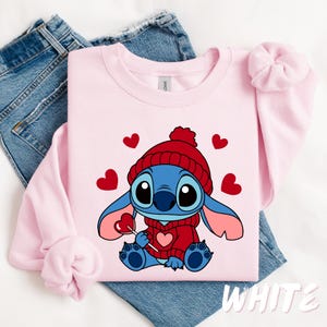 Peut inclure: Sweat-shirt rose avec un personnage bleu Stitch portant un chapeau rouge et tenant une sucette rouge. Le personnage est entour&eacute; de c&oelig;urs rouges. Le texte "WHITE" est visible dans le coin inf&eacute;rieur droit de l'image.