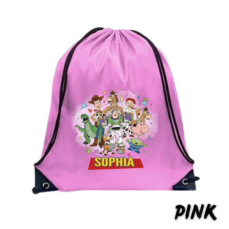 May include: Pink drawstring backpack featuring a colorful graphic of Toy Story characters. The bag has black drawstrings and the name "SOPHIA" in a red banner. The word "PINK" is printed in black at the bottom.