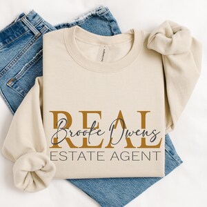 May include: A beige crewneck sweatshirt featuring the text REAL ESTATE AGENT in gold and black lettering, with the name Brooke Owens written in a black cursive script across the center. The sweatshirt is layered over a pair of blue denim jeans.